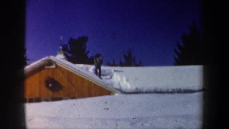 1962 New Hampshire 8mm frame showing a person on a snowy wooden building roof, likely clearing snow under a deep blue sky.