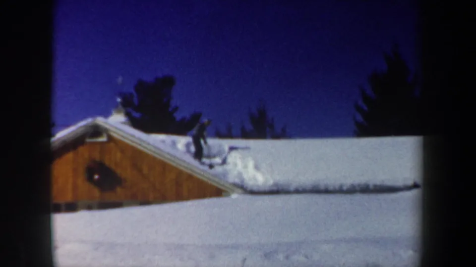 A rooftop figure appears to work in snow on a wooden building in this 1962 New Hampshire winter home-movie frame.