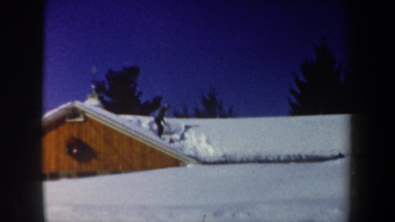 1962 New Hampshire 8mm frame showing a bundled person clearing snow from a cabin roof under a deep blue sky.