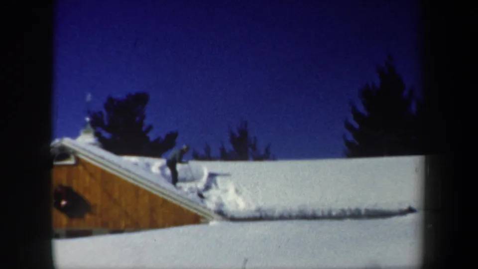 1962 New Hampshire home-movie frame of a person clearing snow from a cabin roof, with a wreath on the wooden wall.