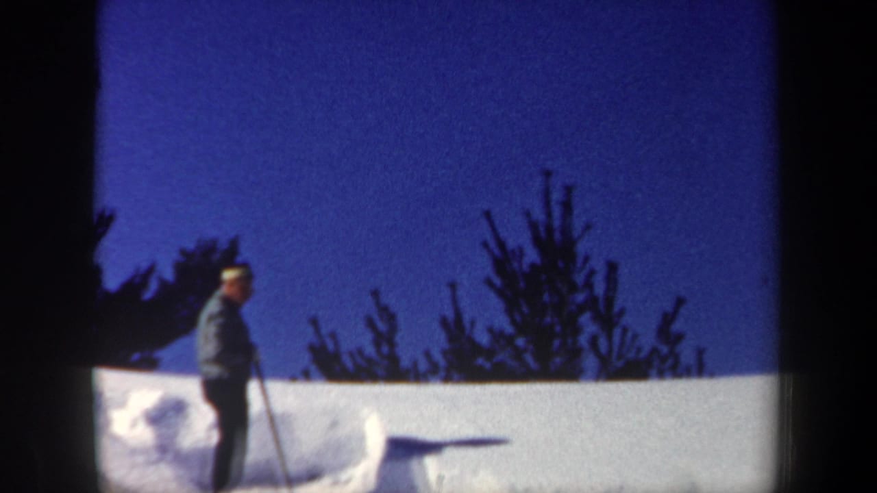 1962 New Hampshire home-movie frame showing a man with a shovel near deep snow and evergreen silhouettes.