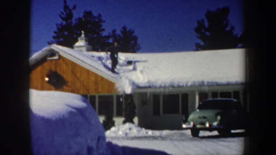 1962 New Hampshire home-movie frame showing a snowy residential roof, wood facade, driveway, and parked green car.