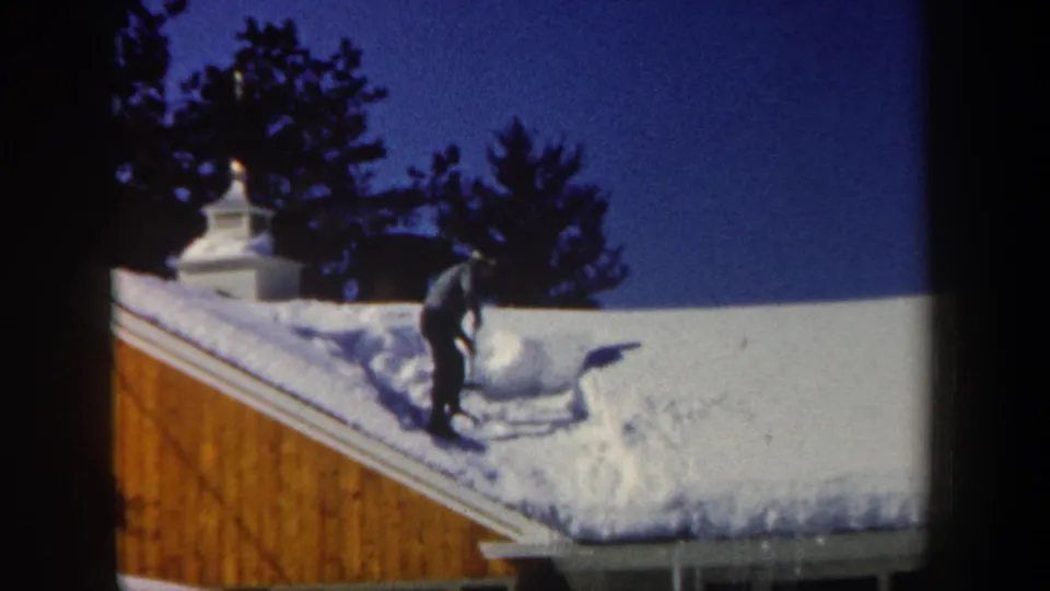 1962 New Hampshire frame of a man clearing deep snow from a sloped residential roof near a chimney.