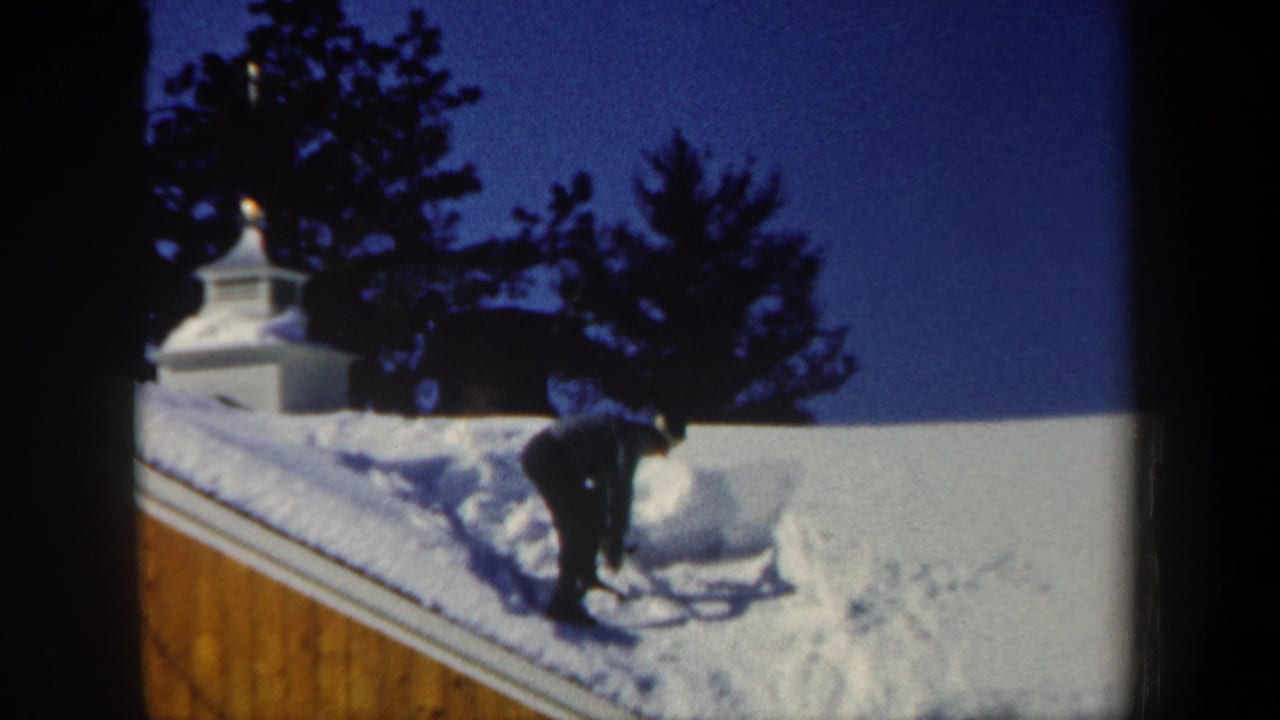 Man bends over with a shovel on a snow-covered roof beside a white cupola and dark trees.