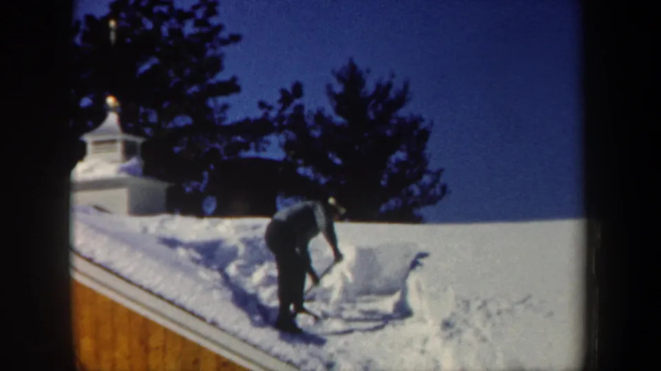 A man in dark winter clothing bends into the task of moving thick roof snow in 1962 New Hampshire.
