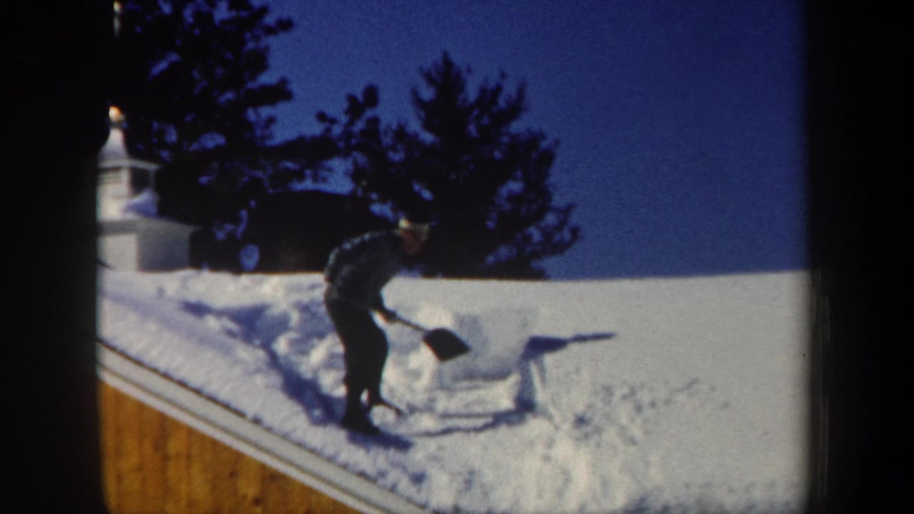 Man shoveling deep snow from a wooden house roof under a blue sky.