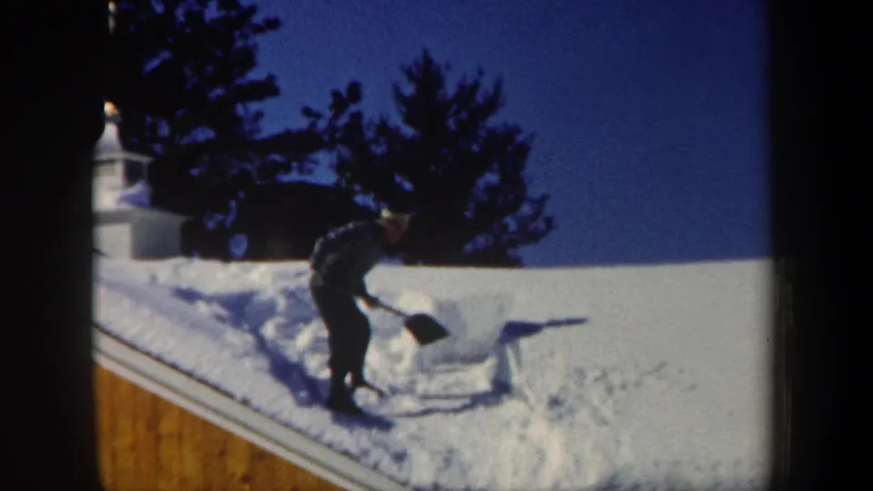 1962 New Hampshire home-movie frame showing a man in dark winter clothing shoveling snow from a wooden house roof.