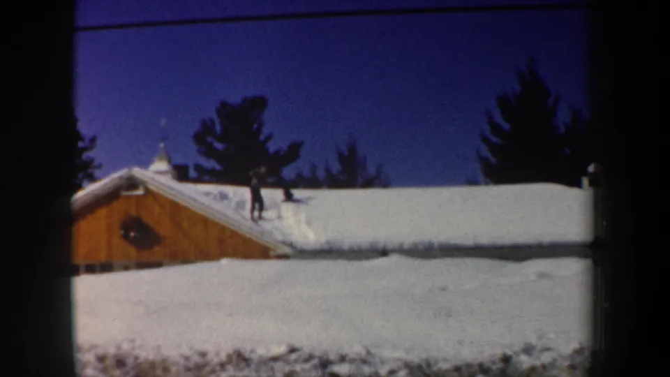 1962 New Hampshire home-movie view of a man on a white roof above a wooden house, with dark evergreens behind.