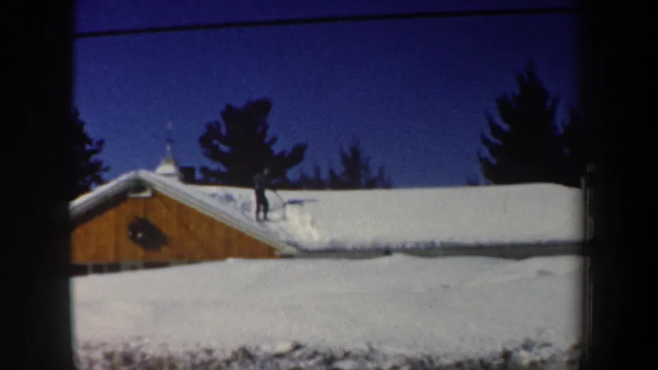 1962 New Hampshire frame showing a distant man on a snowy house roof, with roof snow and evergreen silhouettes in the background.