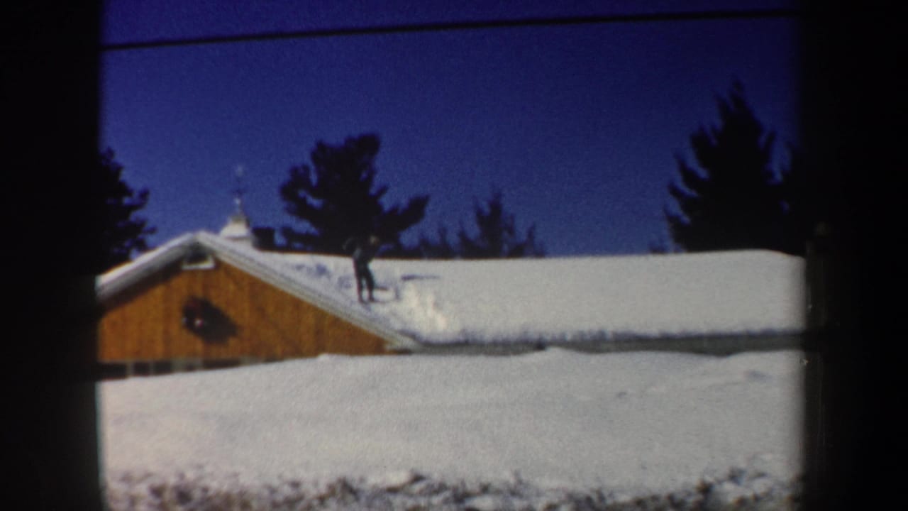 1962 New Hampshire home-movie frame of a man standing on a snowy roof beside a wooden building and evergreen trees.