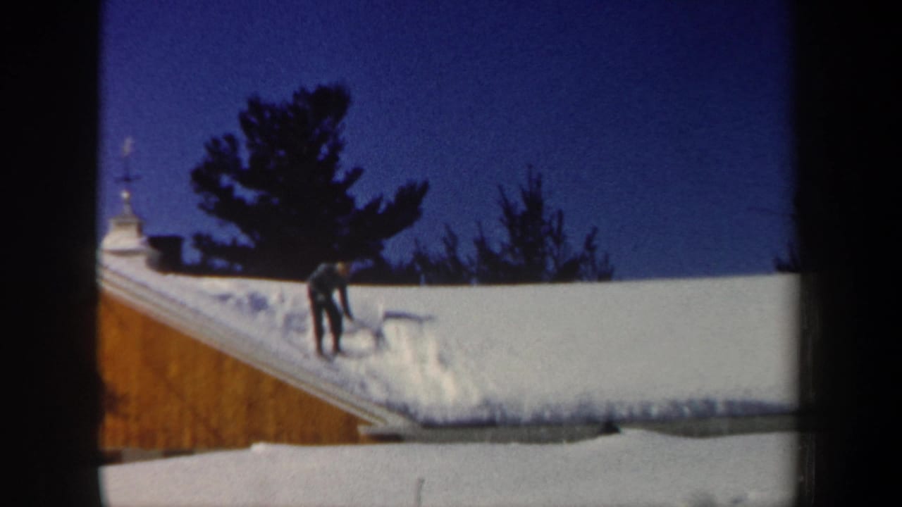 Man bends with a shovel on a snowy roof beside a wooden gable.