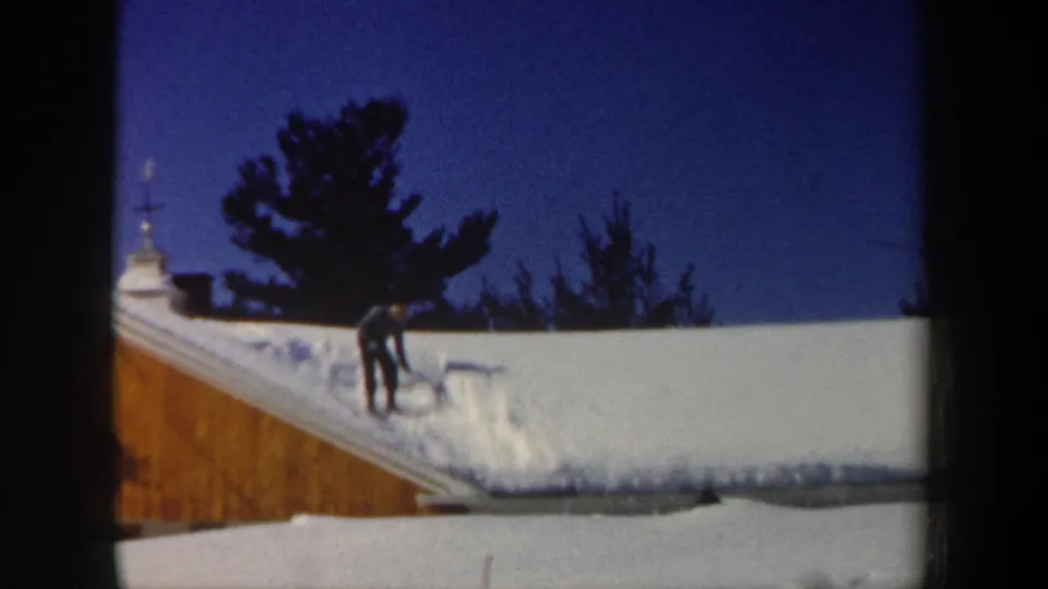 1962 New Hampshire frame showing a man using a shovel on a snow-covered roof with trees and blue sky behind him.