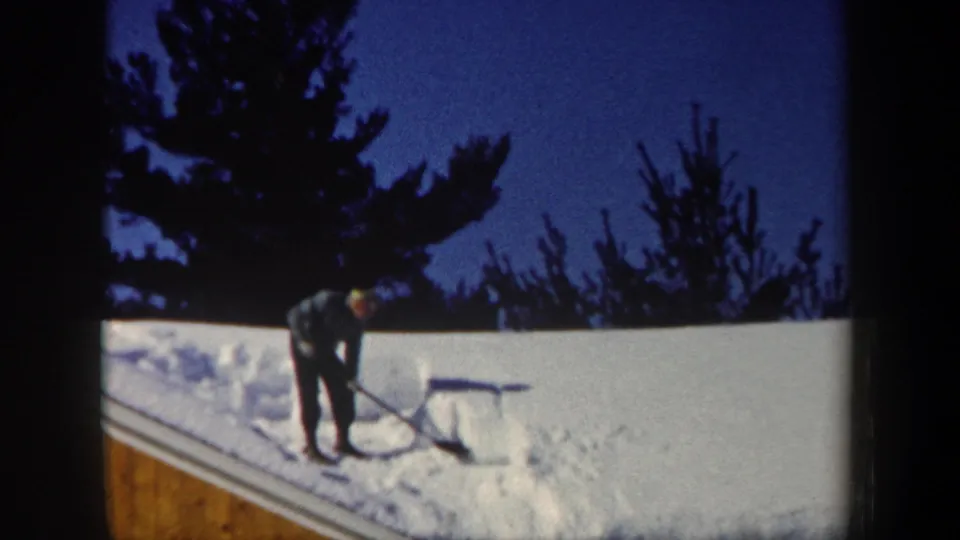 1962 New Hampshire home-movie still with a man in dark winter clothes shoveling roof snow near evergreen trees.
