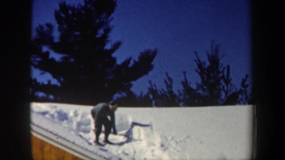 1962 New Hampshire winter frame of a man clearing snow from a roof, with a wooden building edge and dark evergreens visible.