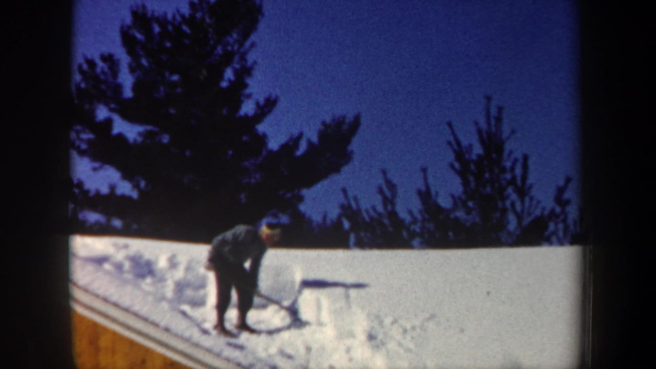 1962 New Hampshire home-movie frame showing a man beginning to clear snow from a sloped house roof with evergreens behind him.