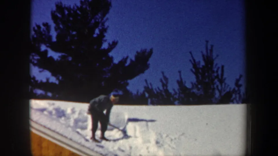 1962 New Hampshire home-movie frame showing a man beginning to clear snow from a sloped house roof with evergreens behind him.