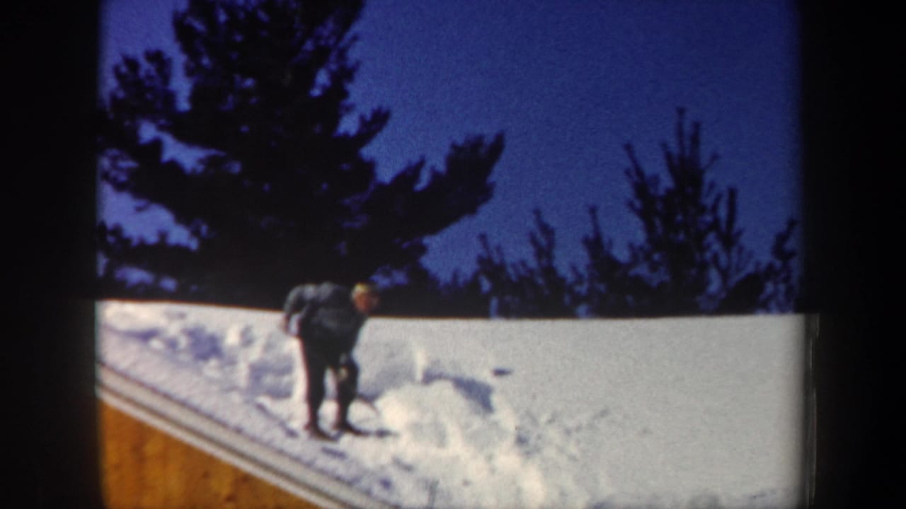 Man bends over with a shovel while clearing deep snow from a roof.