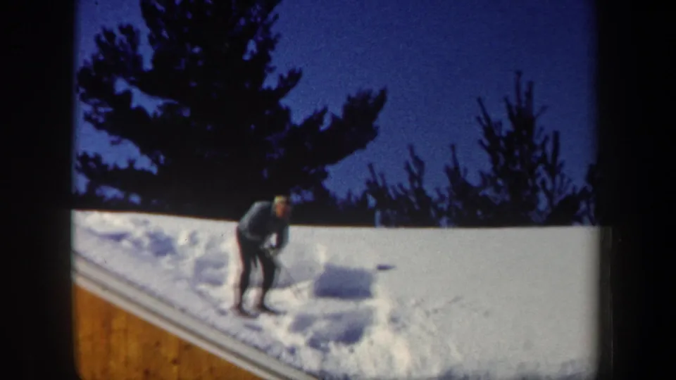1962 New Hampshire home-movie frame showing progress as a man shovels roof snow beside a cleared patch and snow pile.