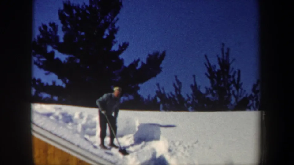 1962 New Hampshire frame showing a man standing with a shovel near a large mass of snow on a roof, with trees in the background.