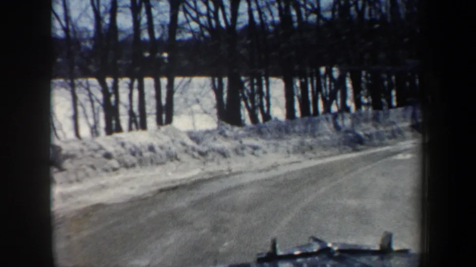 1962 New Hampshire road-view frame from a car moving through snowbanks and leafless roadside woods.
