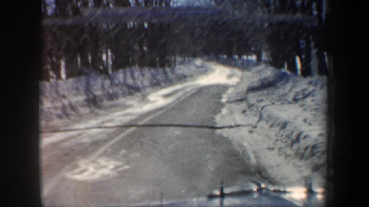 Icy paved road curving between snowbanks and leafless trees.