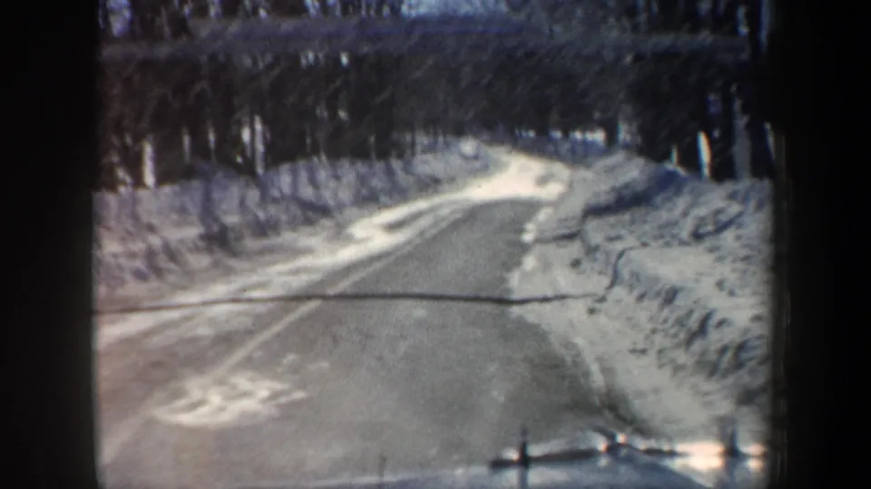 Snowy New Hampshire road in 1962 viewed from a vehicle, with icy pavement, high snowbanks, and leafless trees.