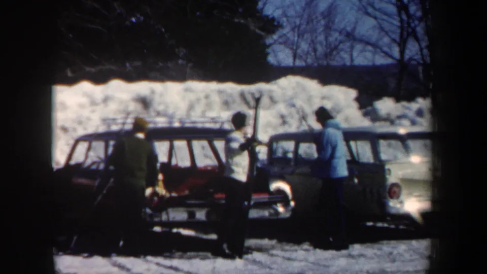 Three people in winter coats unload or handle gear beside vintage station wagons in snowy New Hampshire.