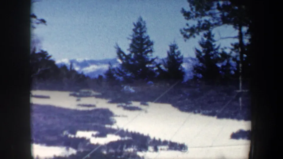 1962 New Hampshire home-movie frame looking past dark evergreens toward frozen water and a wooded valley.