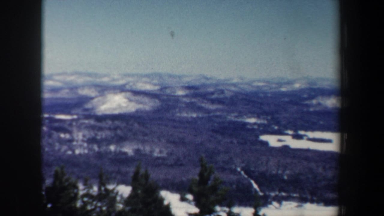 Distant winter mountains and wooded valley with snow patches under a pale sky.