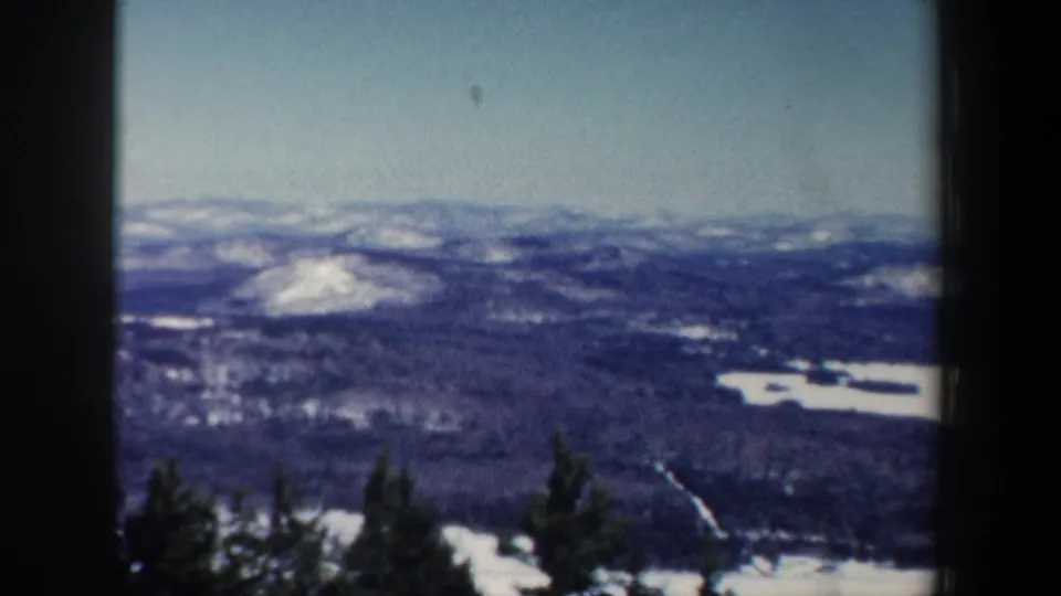 1962 New Hampshire home-movie frame with layered snowy hills, dark woodland, and a possible frozen lake below.