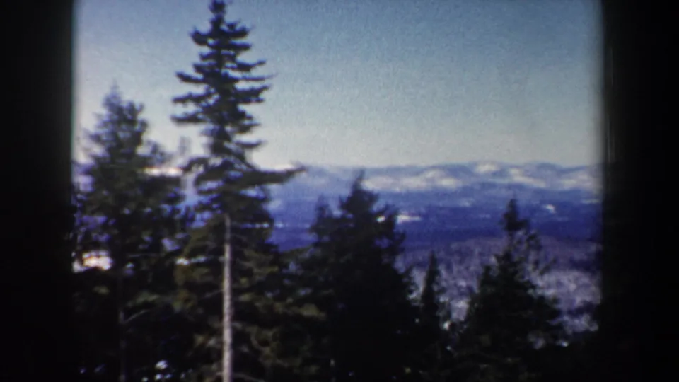 1962 New Hampshire 8mm frame showing tall conifers, a wooded slope, and hazy snow-covered ridges beyond.