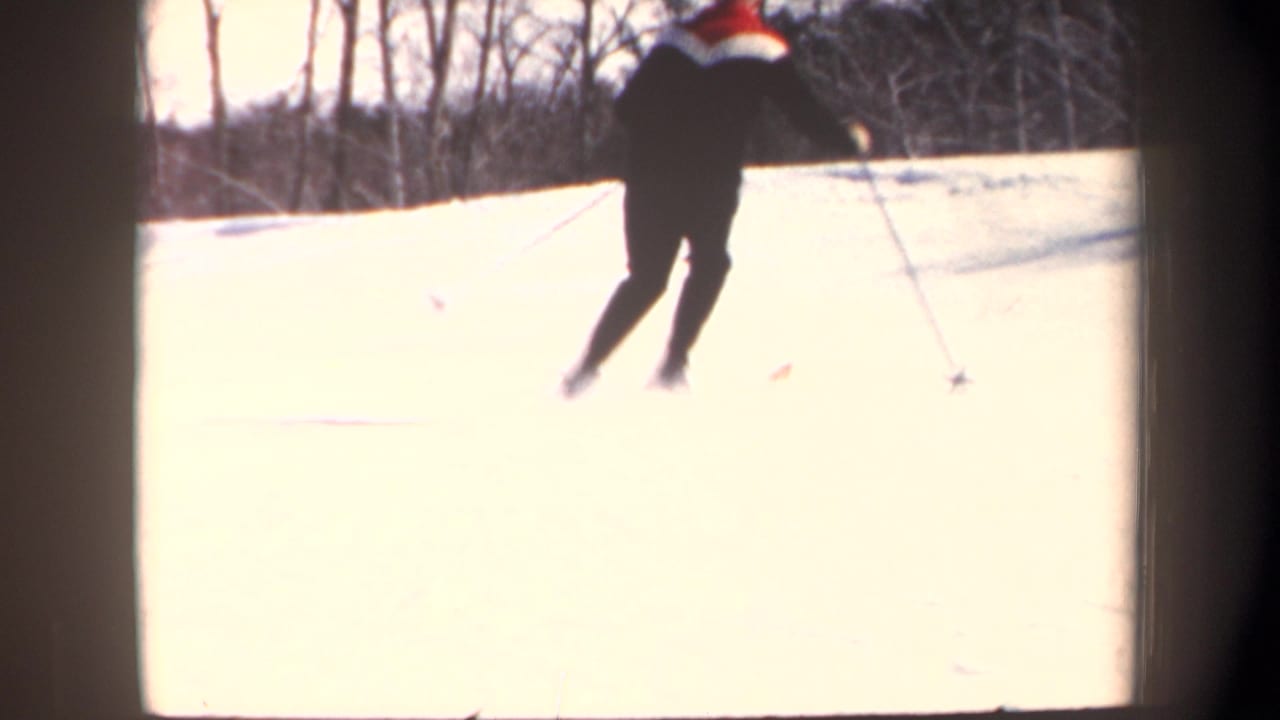 In 1962 New Hampshire home-movie footage, a man in dark winter clothing practices skiing on a snowy hill bordered by bare trees.