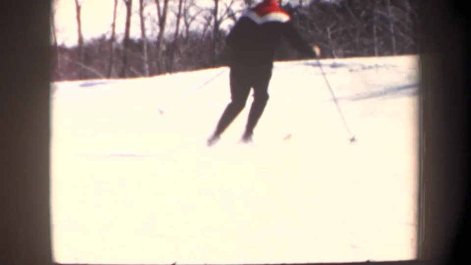 In 1962 New Hampshire home-movie footage, a man in dark winter clothing practices skiing on a snowy hill bordered by bare trees.