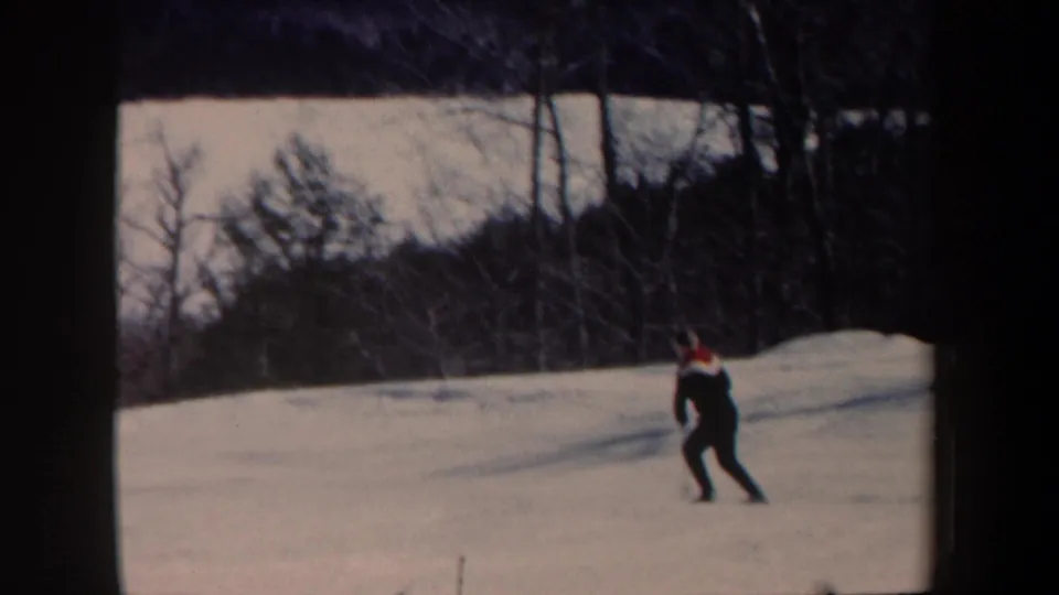 The home-movie frame shows a distant adult skier traversing a gentle snowy hill, with woods and winter trees behind him.
