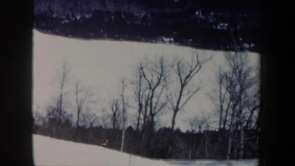 1962 New Hampshire home-movie frame showing a snowy slope, leafless trees, and a distant skier in dark clothing.