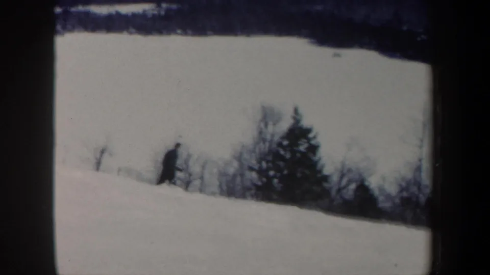 Closer 1962 New Hampshire home-movie view of a lone skier descending beside bare trees and an evergreen on a snow-covered hill.