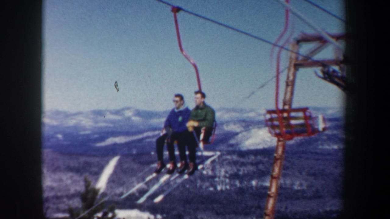 In 1962 New Hampshire home-movie footage, two adult men ride a red chairlift over snowy mountain terrain under a clear sky.