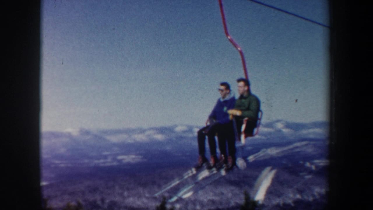 Two men sit on a ski-lift chair suspended above snowy hills and evergreen slopes.