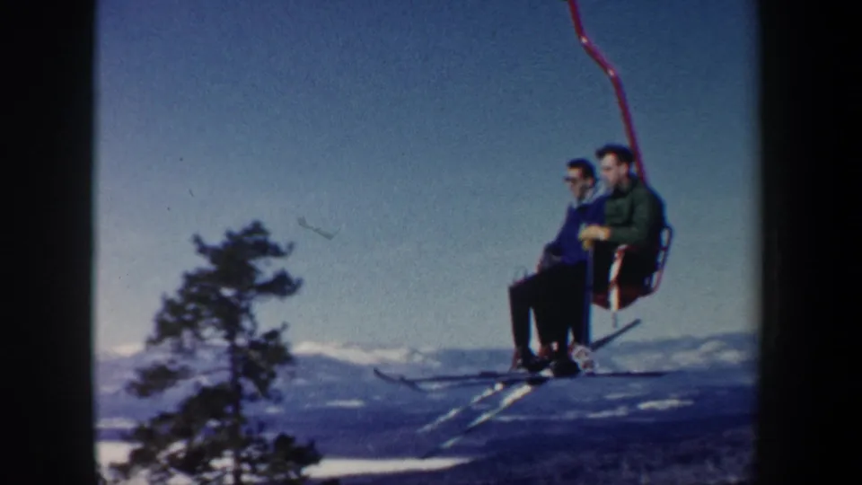 The 8mm frame shows two adult men on a red ski lift passing above an evergreen, with New Hampshire winter hills in the distance.