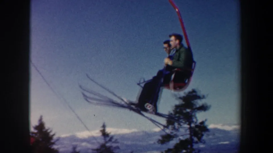 Two adult skiers on a red chairlift move past evergreen trees, their skis visible against the snowy New Hampshire mountain backdrop.
