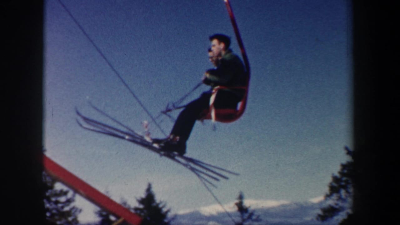 Skier seated on a red chairlift with long skis against a blue sky.