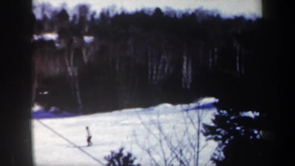 1962 New Hampshire 8mm frame showing a distant lone skier on a snowy run below a wooded hillside.