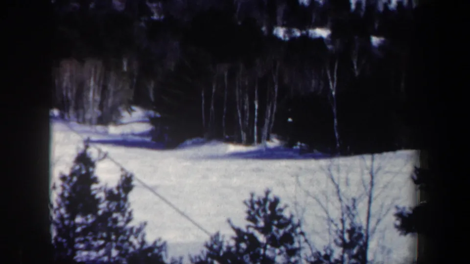 1962 New Hampshire winter home-movie frame with snow, forest, and a diagonal cable line across the slope.