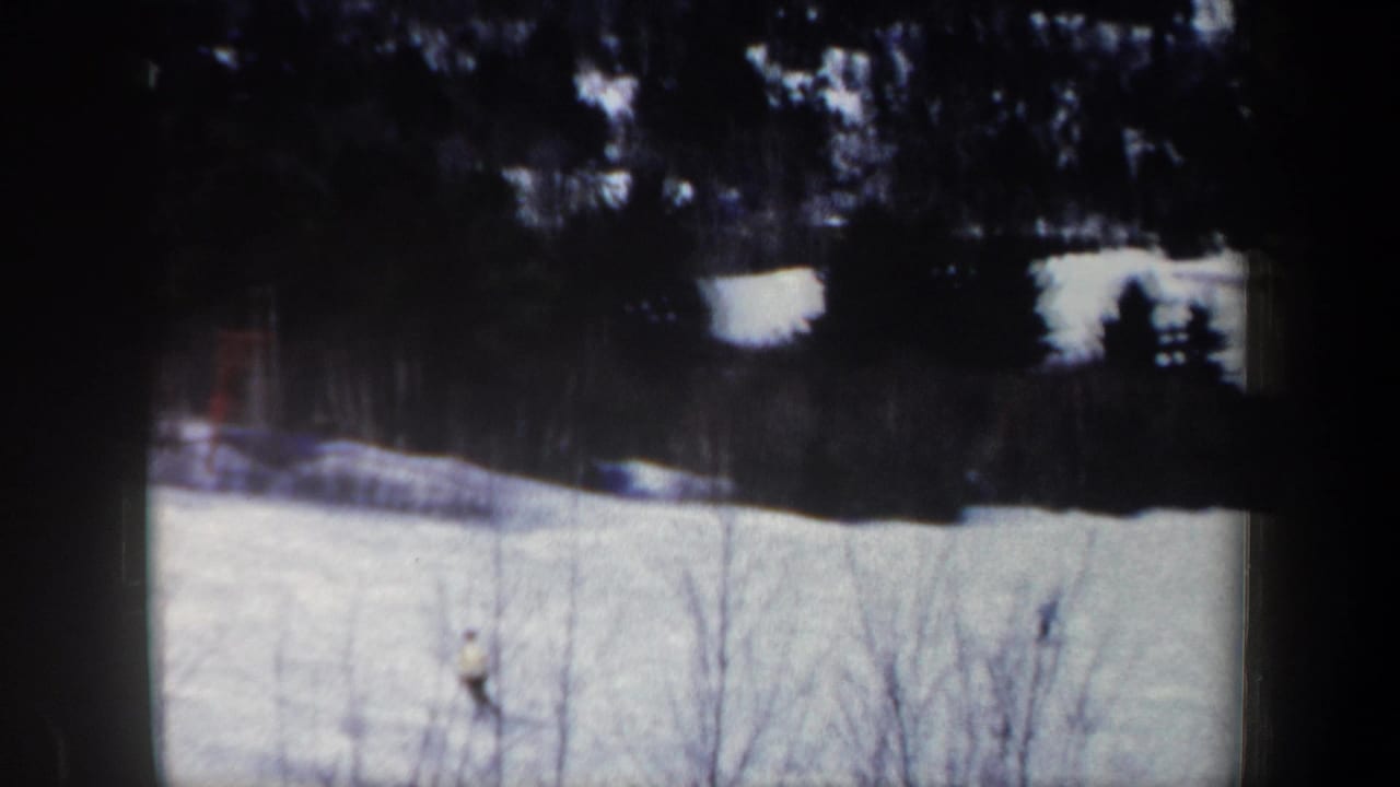 1962 New Hampshire 8mm frame showing a distant skier in pale winter clothing moving across a snowfield below dark trees.