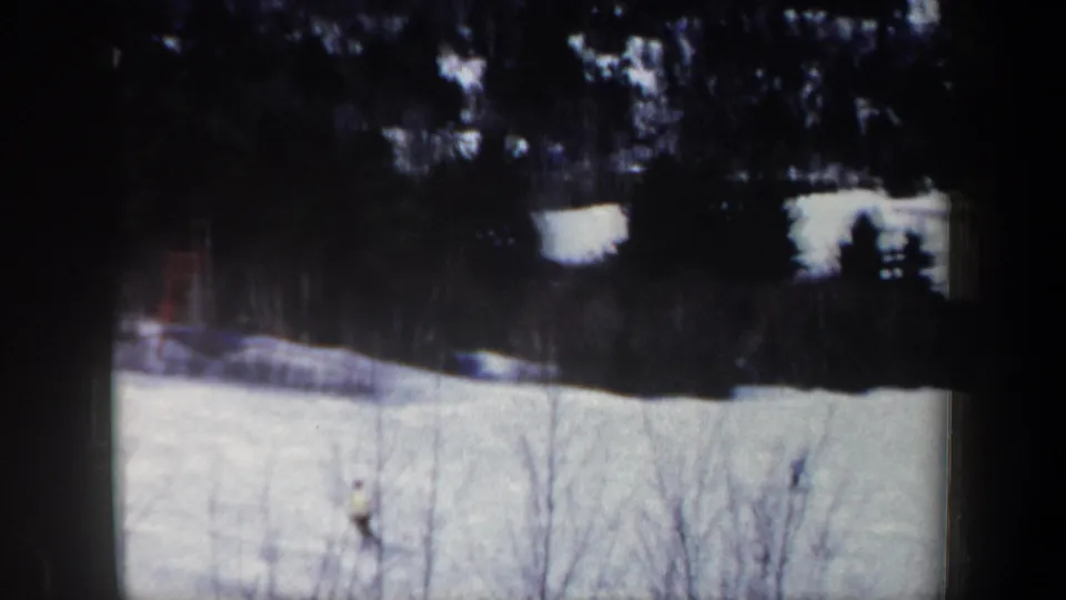 1962 New Hampshire 8mm frame showing a distant skier in pale winter clothing moving across a snowfield below dark trees.