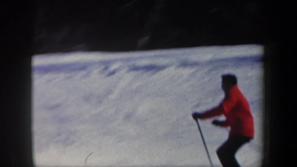 1962 New Hampshire 8mm frame showing an adult skier in a red jacket moving across snow with ski poles near a wooded edge.