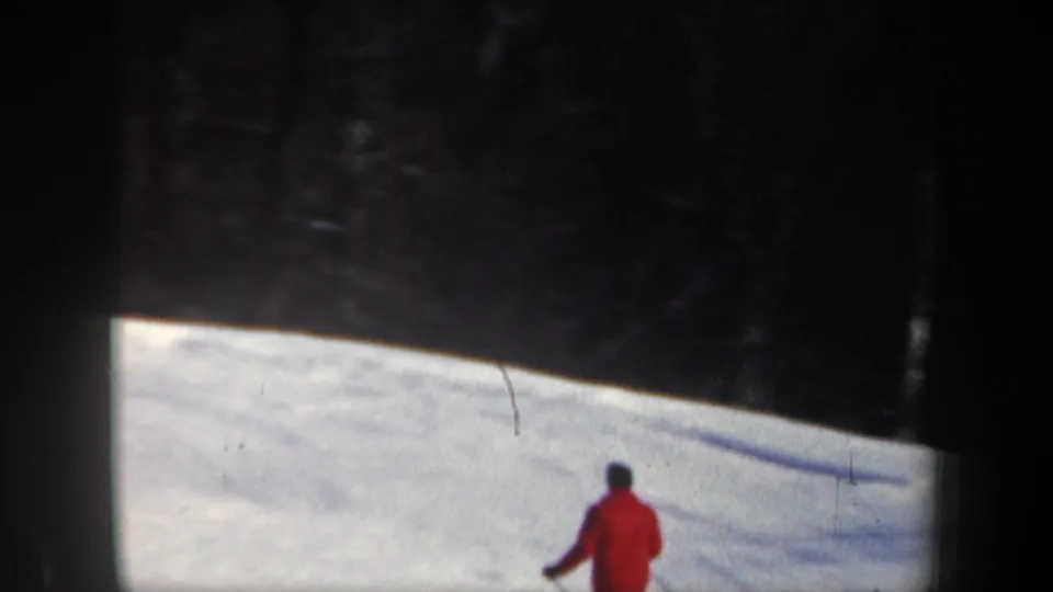 1962 New Hampshire 8mm frame showing a distant red-jacketed skier on a bright snowfield below dark wooded shadow.