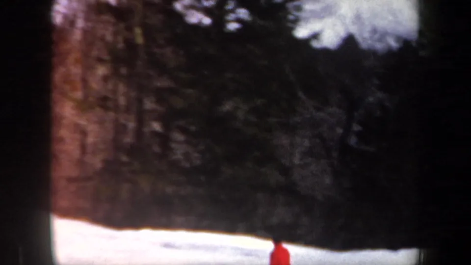 1962 New Hampshire home-movie frame with a partially framed red-jacketed skier below a dark evergreen backdrop and snowbank.