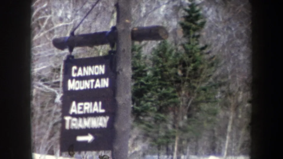 1962 8mm frame showing a Cannon Mountain Aerial Tramway sign with white lettering and arrow in a snowy forest setting.