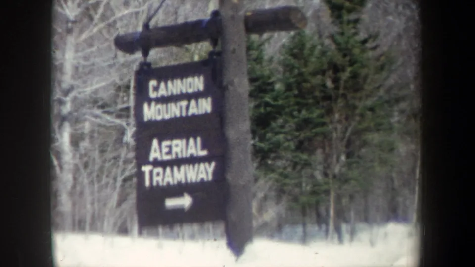 The sign for Cannon Mountain Aerial Tramway appears closer in frame, mounted on a wooden post among snow, evergreens, and bare trees.